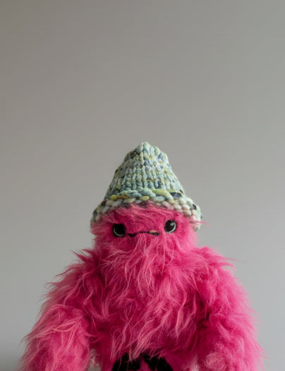Pink fluffy creature with a colorful beanie on a neutral background