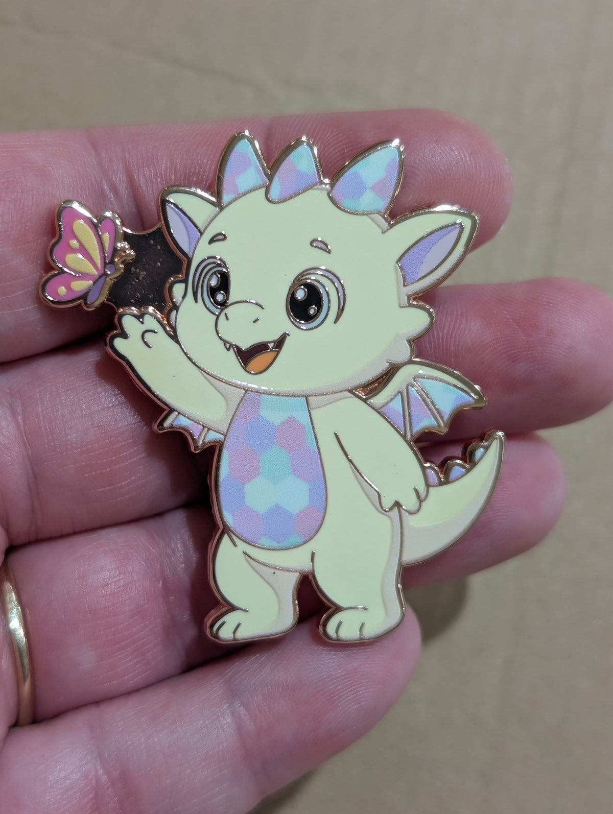 Opaliath enamel pin: a buttery yellow baby dragon with opal pastel scales waving at a butterfly